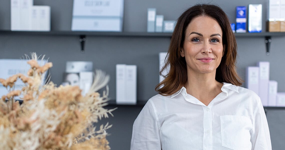 žena dívající se do fotoaparátu, stojí v interiéru a za sebou má vystavené produkty Oslo Skin Lab, Michelle Ottendal 